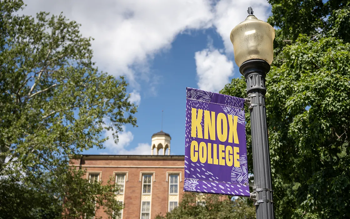 A photo of a purple light pole banner with the words "Knox College" in yellow. The historic brick building, Old Main, and green trees are in the background.