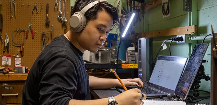 A student in headphones sits in front of a laptop and takes notes on a pad of paper.