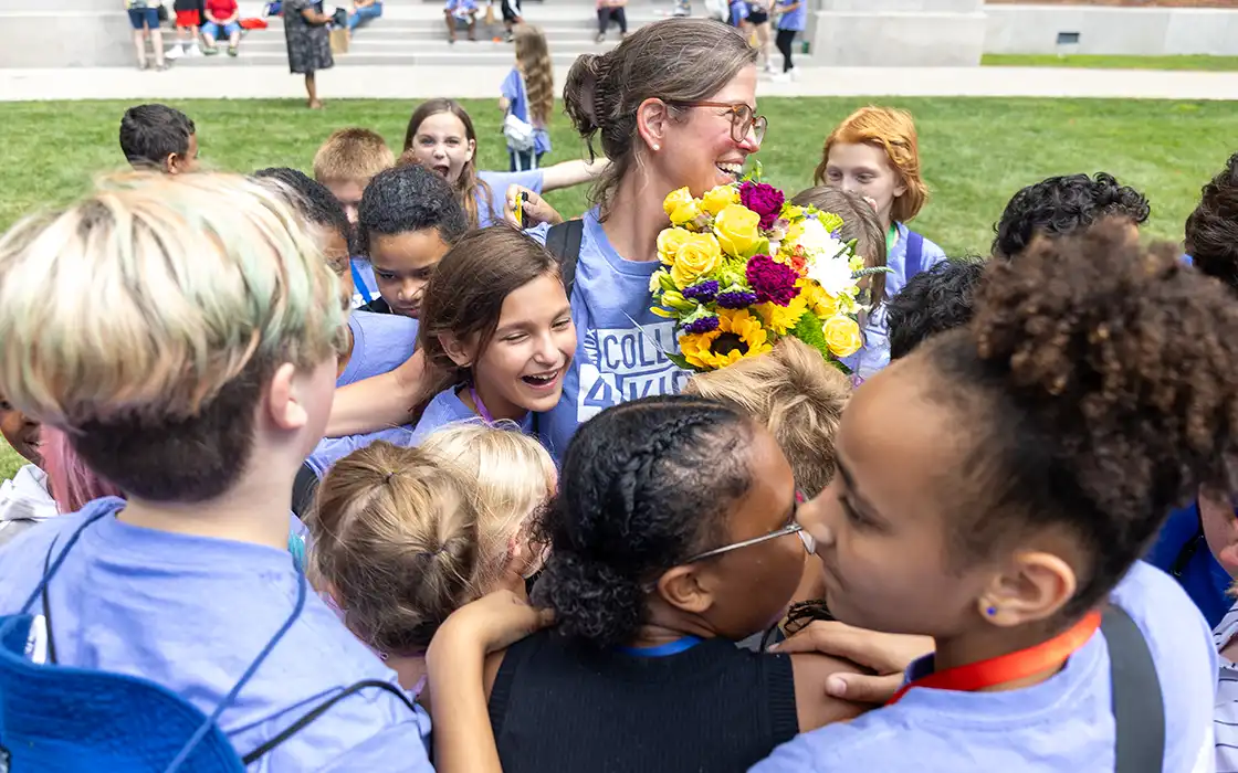 A group of elementary school students surround a Knox College 4 Kids tteacher, who holds a big bouquet of flowers.