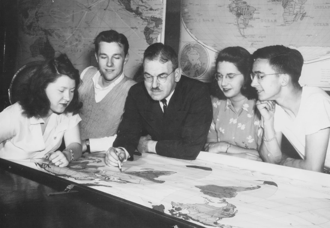 A black and white photo of four Knox students gather around a Knox professor, who is pointing to a map on a table.