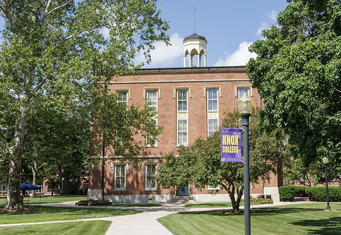 A photo of Knox College's Old Main on a sunny day. A light pole with a banner that says "Open Arms" is in the foreground.