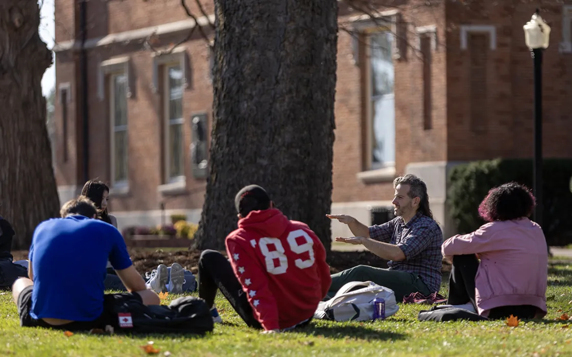 A professor and students hold a class discussion while sitting in a circle on the campus lawn.