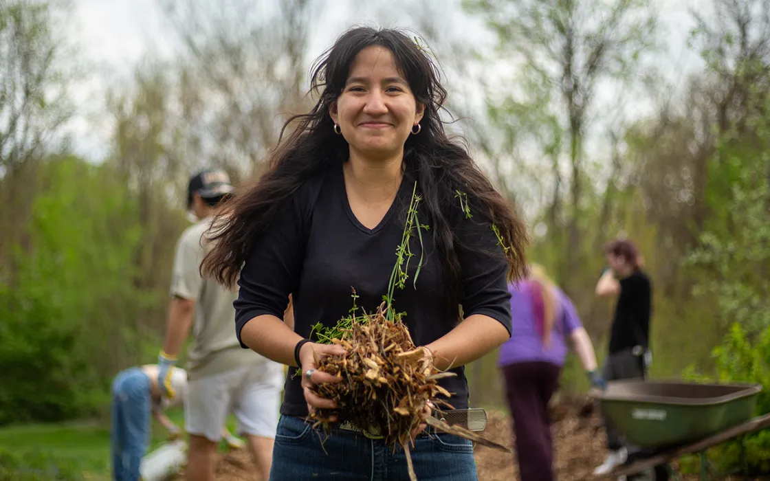 A smiling Knox student holds falls leaves in her hands as other students rake leaves in the background.