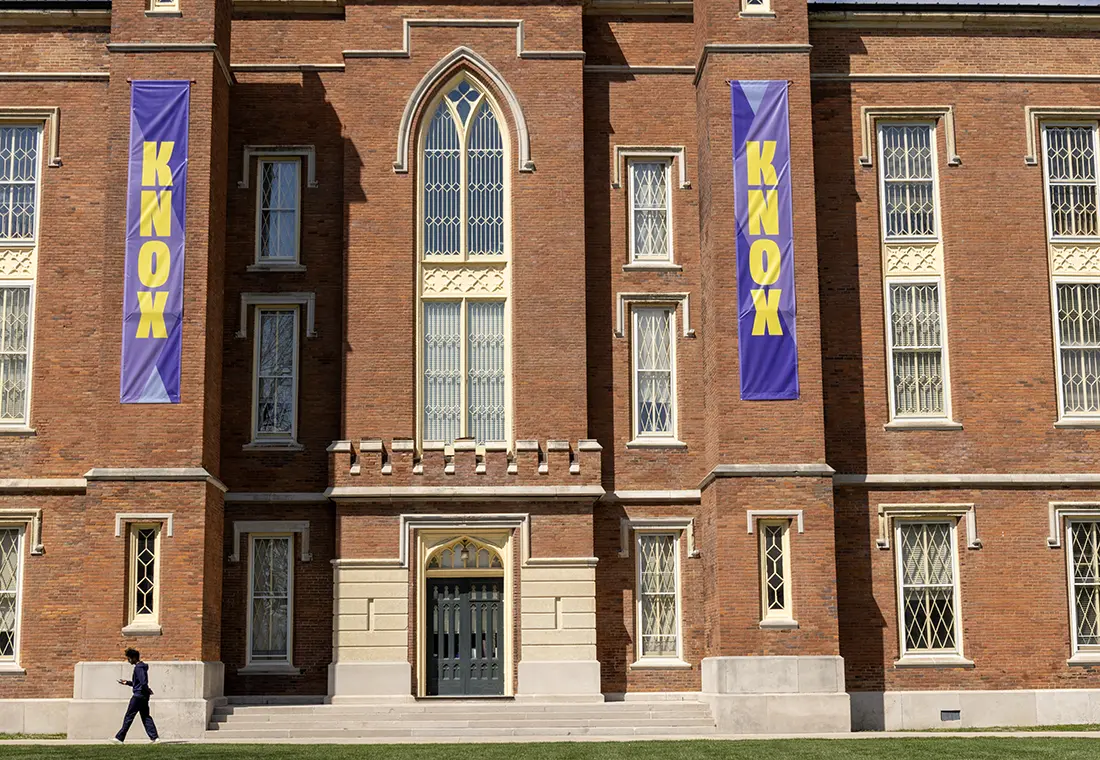 A students is walking in front of the south side of Old Main. Two purples banners that say "Knox" in yellow hang on either side of the building.