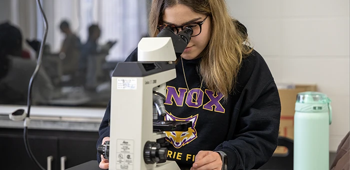 A student wearing a Knox shirt looks into a microscope