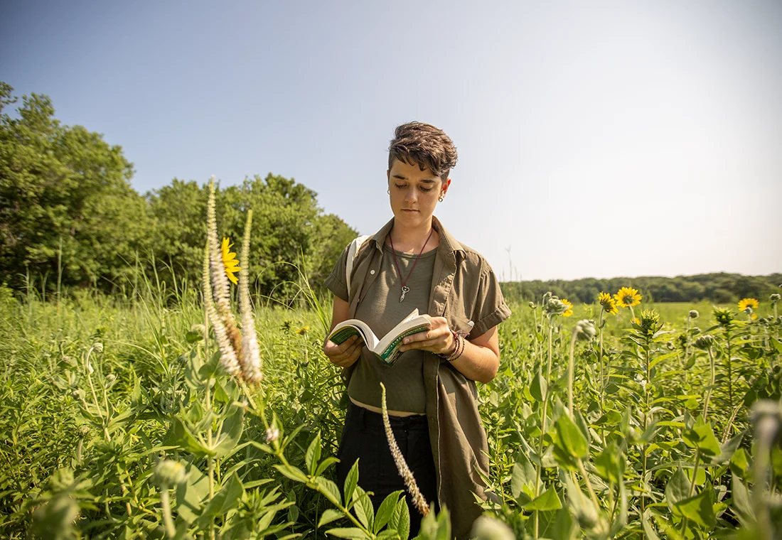 A student stands in an open green field writing in a notebook.
