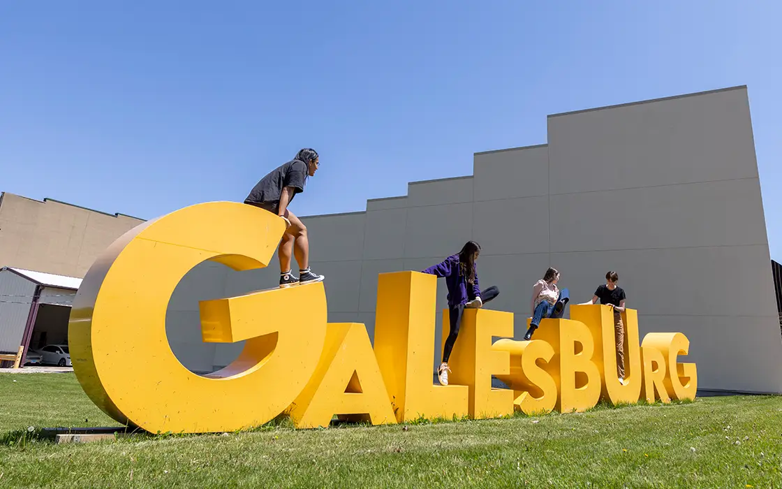 Four Knox students sit on top of a large yellow Galesburg sign in a field.