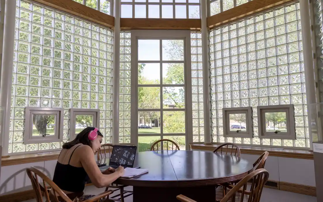 A student sits at a table in the library with a laptop, and is taking notes.