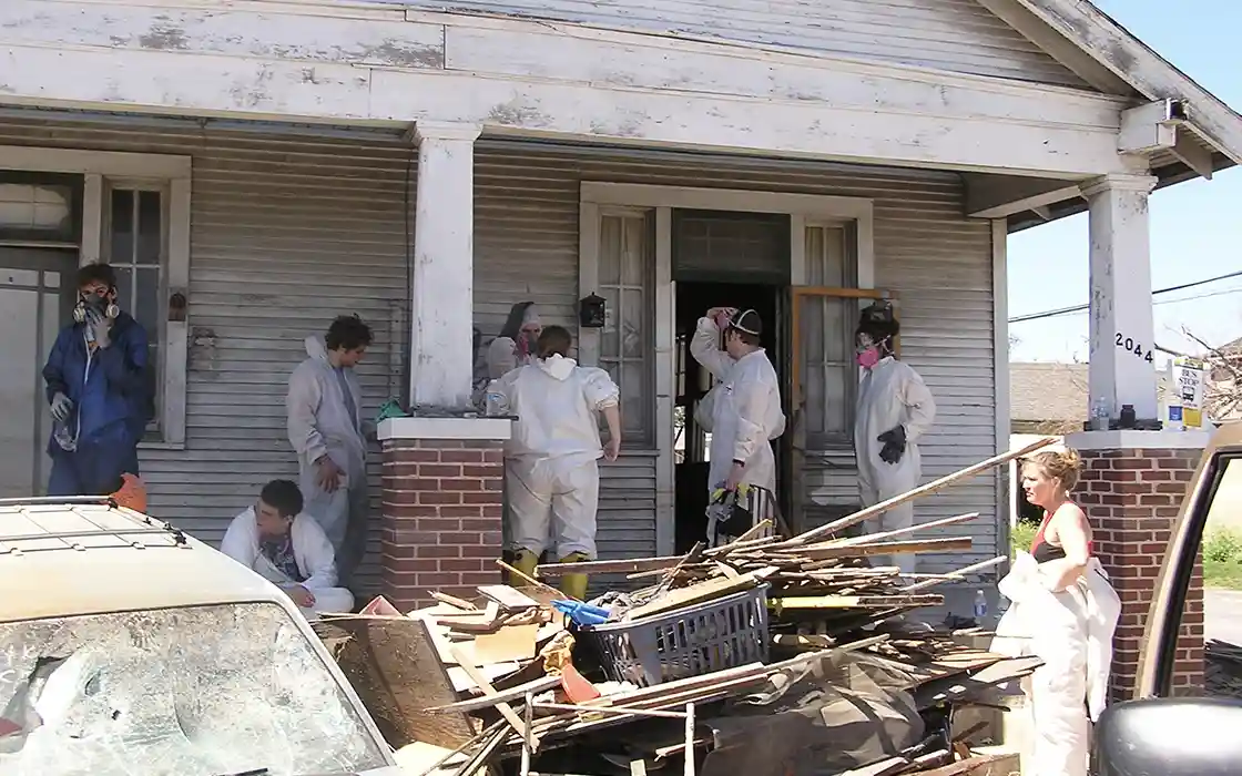 A group of students wearing cleaning gear stands on the front steps of a house, next to materials that had been cleaned up.