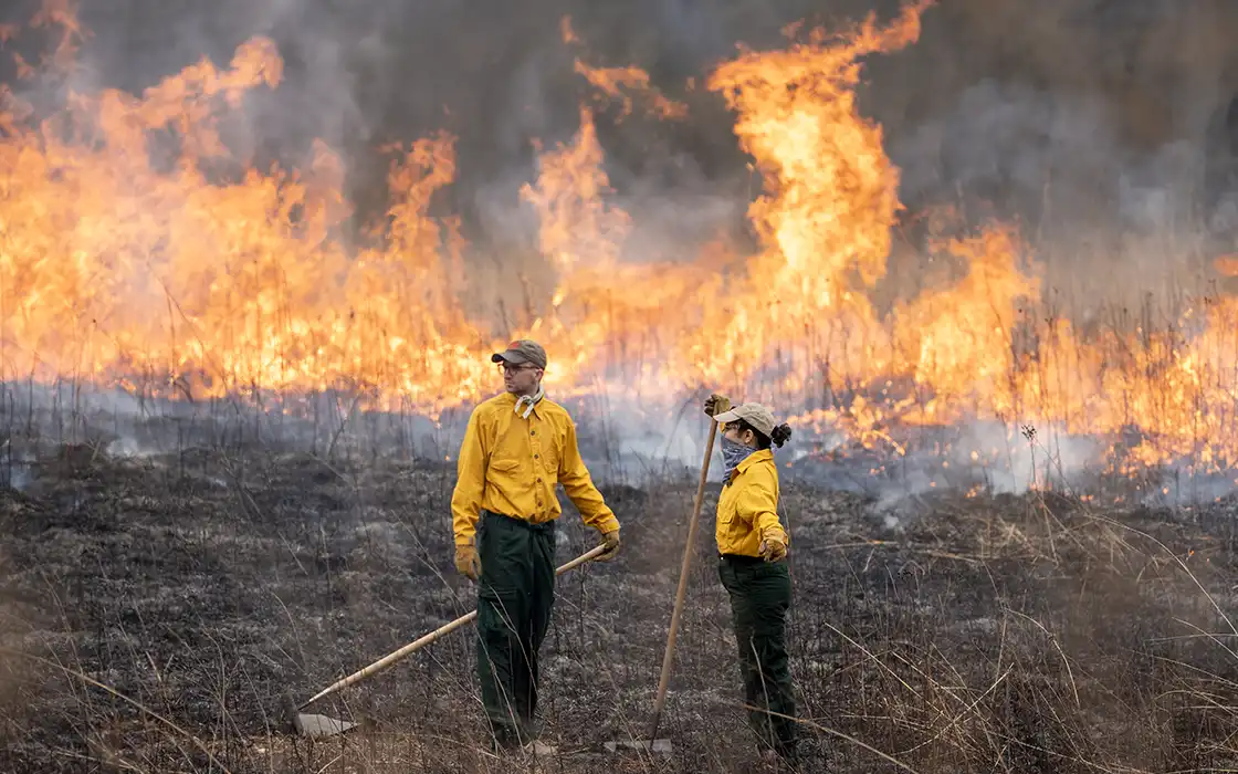 Two students stand in the prairie with a fire burning in the background.