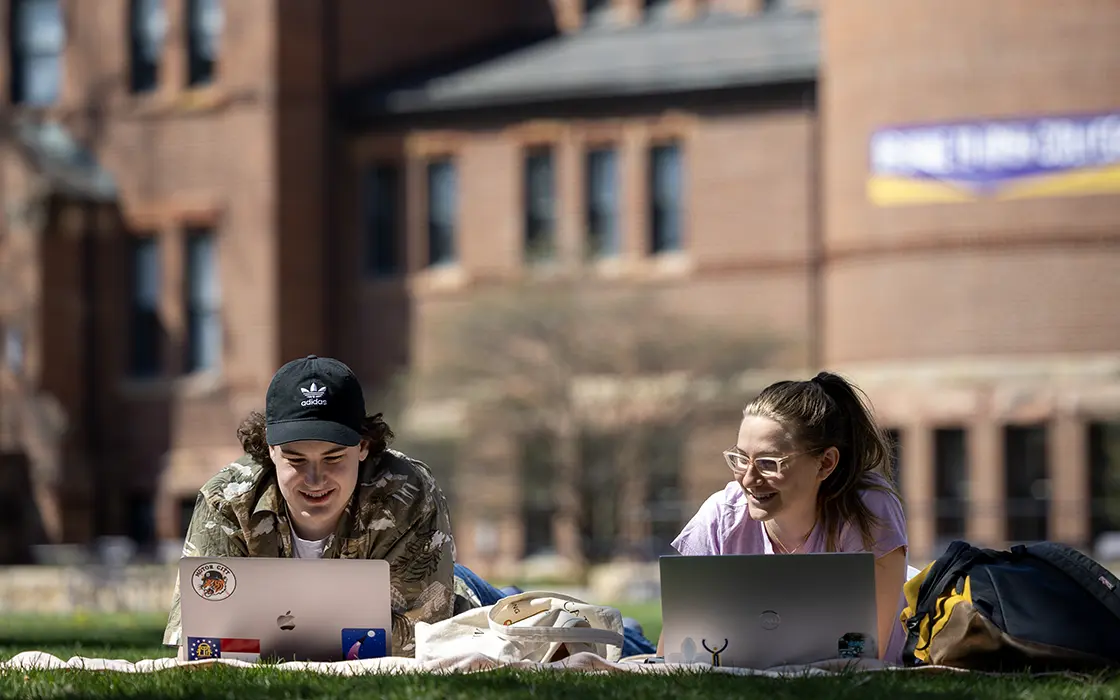 Two students lay out on the grass with their laptops