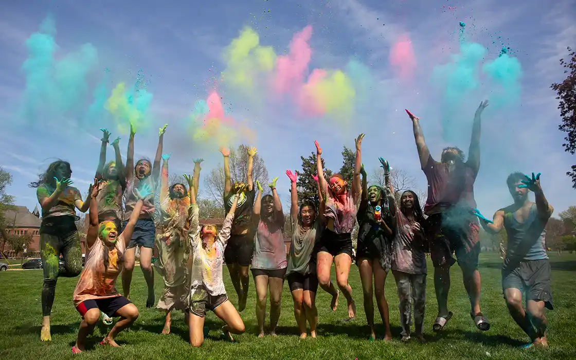 A group of Knox students are throwing colored powder in the air at a Holi celebration.