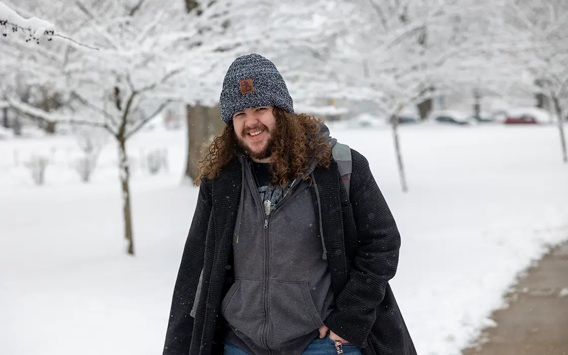 A student walks across a snow-covered campus in a hoodie and parka.