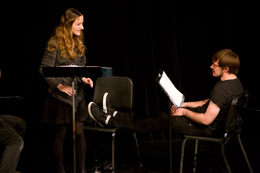 Two students on a dark and bare theatre stage. One reads to the other from a script.