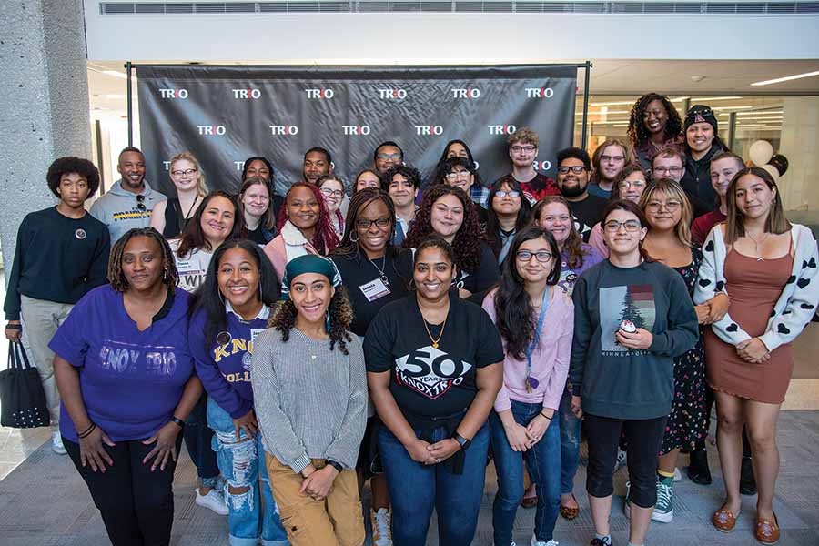 A group of TRIO students and alums take a photo in front of a black backdrop with TRIO printed across it. 