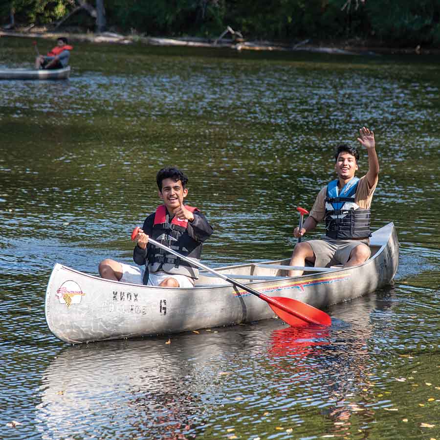 Two students in a canoe on a lake at Green Oaks. One student has his thumb up and the other student is waving to the camera. 