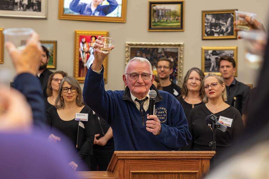 Harley Knosher standing behind a podium with people in front of him and behind him. He holds up a glass during a toast.