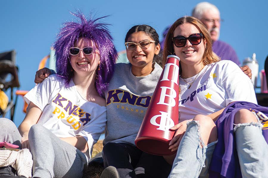 Three students sitting on the hill at the Knosher Bowl during the Homecoming game. One has a purple wig on.