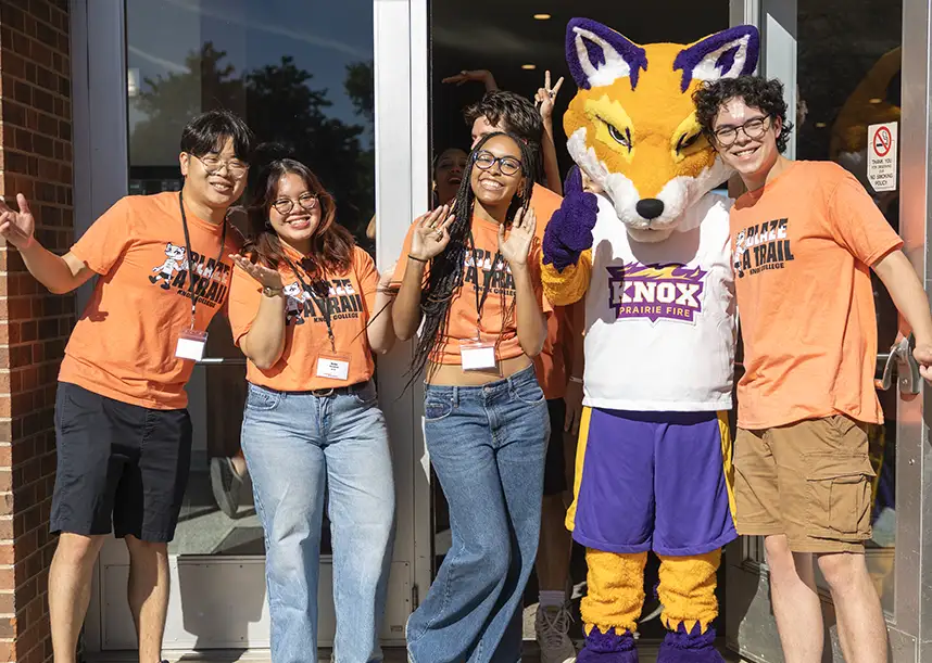 A group of smiling students surround the Knox College mascot, Blaze, a purple, gold, and white fox.