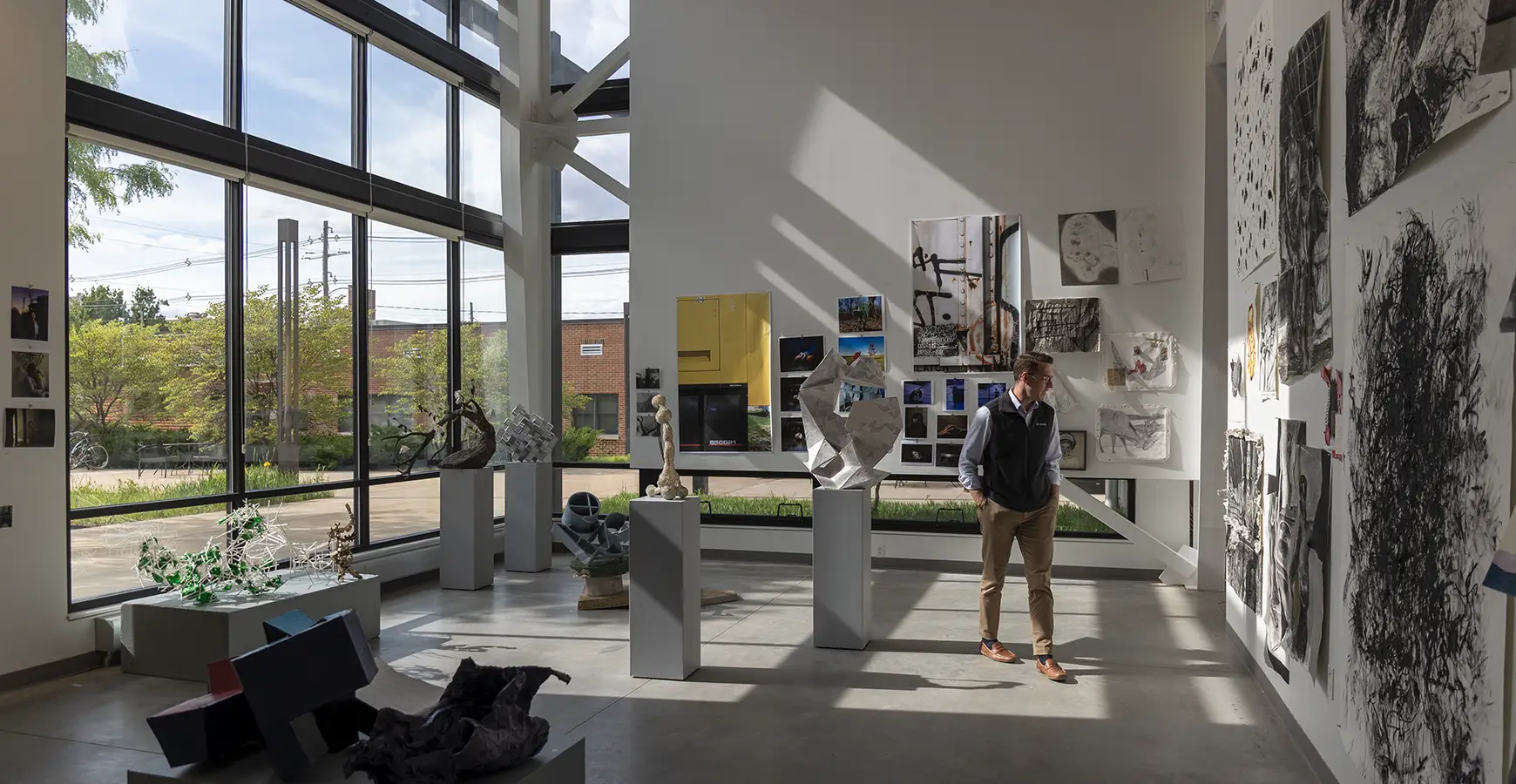 Professor Thomas Bell regards art hung on a wall in the Whitcomb Art Center. Sunlight streams in through the windows behind him and shines on the art.