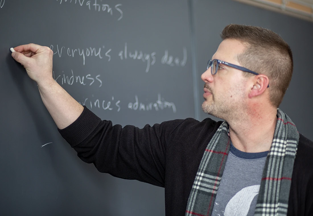 During a fiction workshop, a professor writes on a chalkboard.