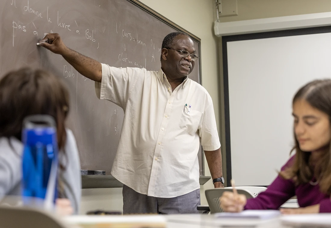 A professor points at a chalkboard where a sentence is written in French, partially visible. Two students are visible in the foreground.