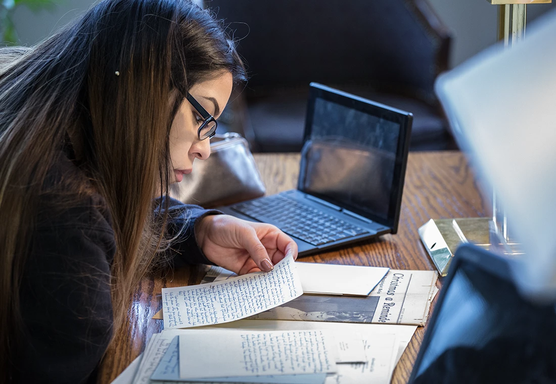 A student leans down to read a handwritten letter. Behind the letter, an old issue of a newspaper is on the table. A laptop computer is open next to the student.