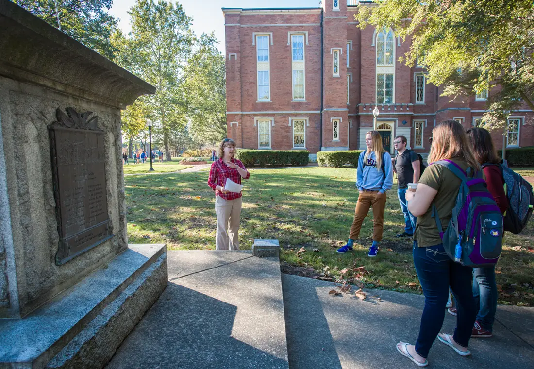 A woman speaks, gesturing toward a monument, while a group of students gathers around and listens. Old Main is behind them.