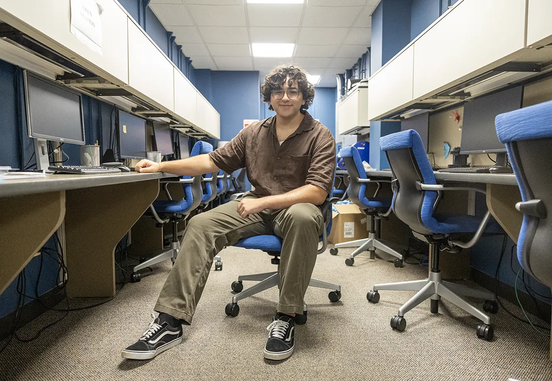 A student sits in the middle of a room surrounded by desks and computers.