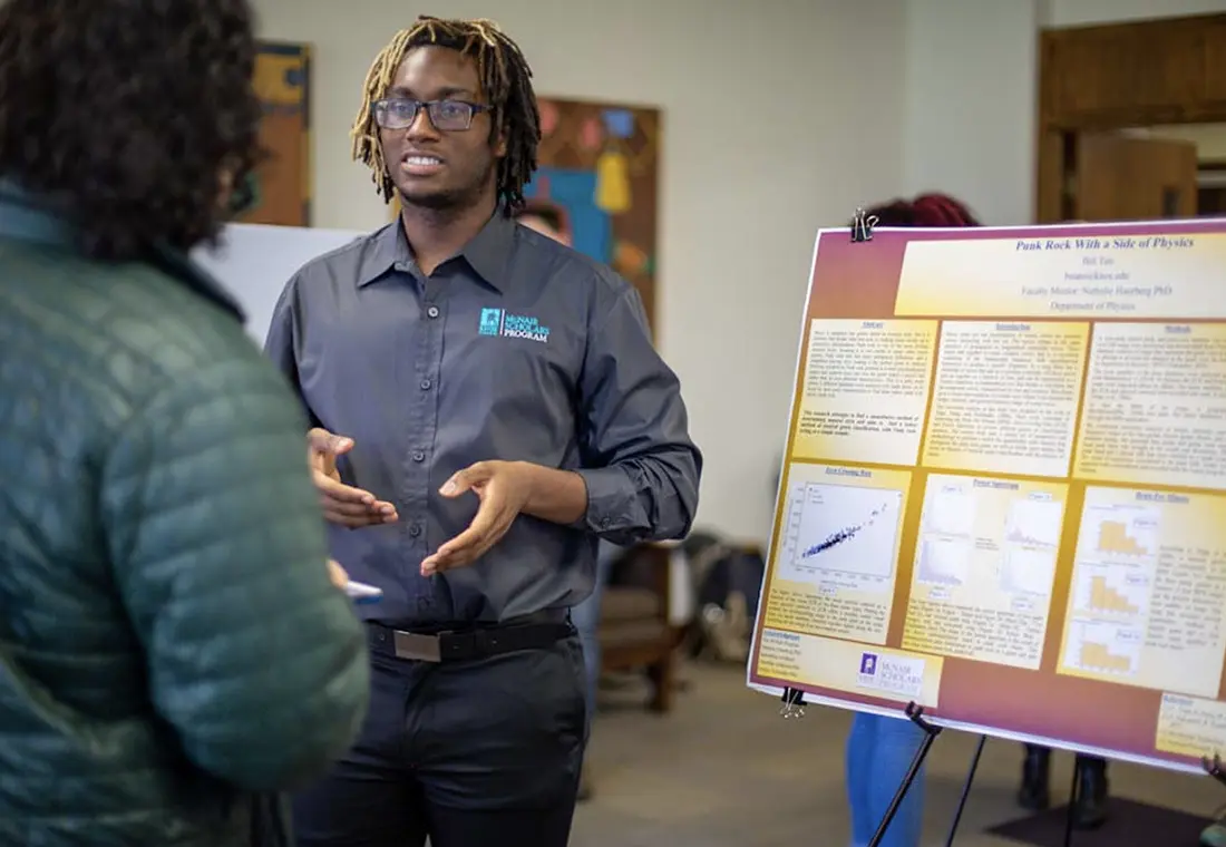 A student presents to a person while standing in front of a poster board.