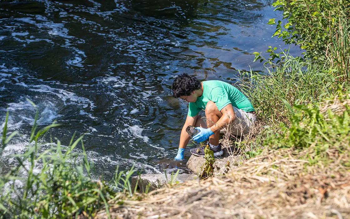 A student sits at the edge of a body of water, collecting a sample in a mason jar.