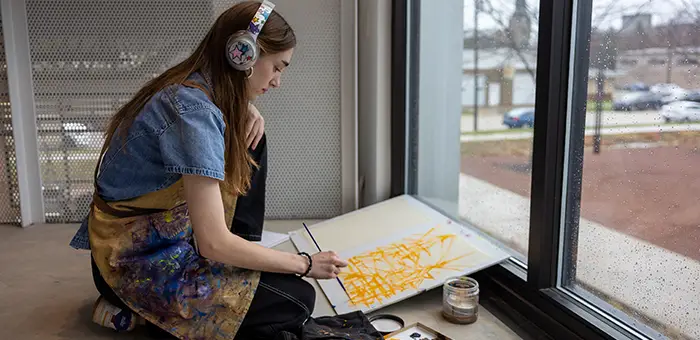 A art student wearing headphones looks out a window while painting.