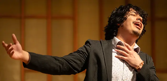 A music student sings on a stage with a hand on their heart and arm outstretched to the side. to the