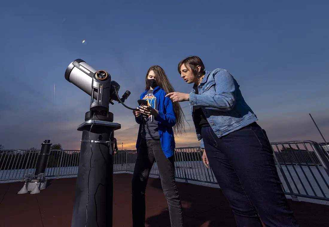A Knox professor and a student standing behind a telescope with the night sky behind them.