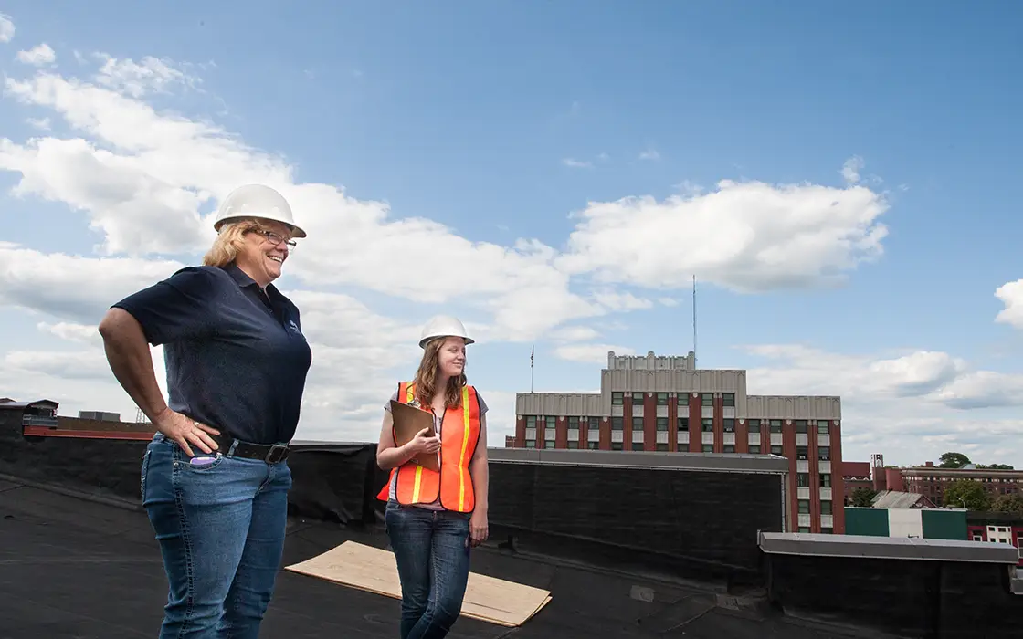 To woman overlook a construction site, one is a Knox student wearing a hard hat and orange vest, the other is a wearing a hard hat with no vest.