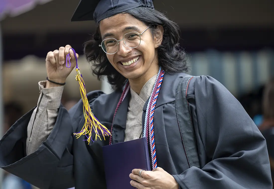 A Knox graduate wearing their cap and gown smiles at the camera while holding up their graduation tassle.