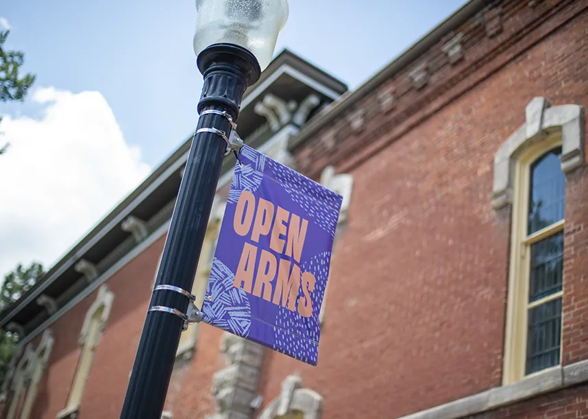 A lightpole purple banner with yellow letters that say "open arms" in front of a brick building.