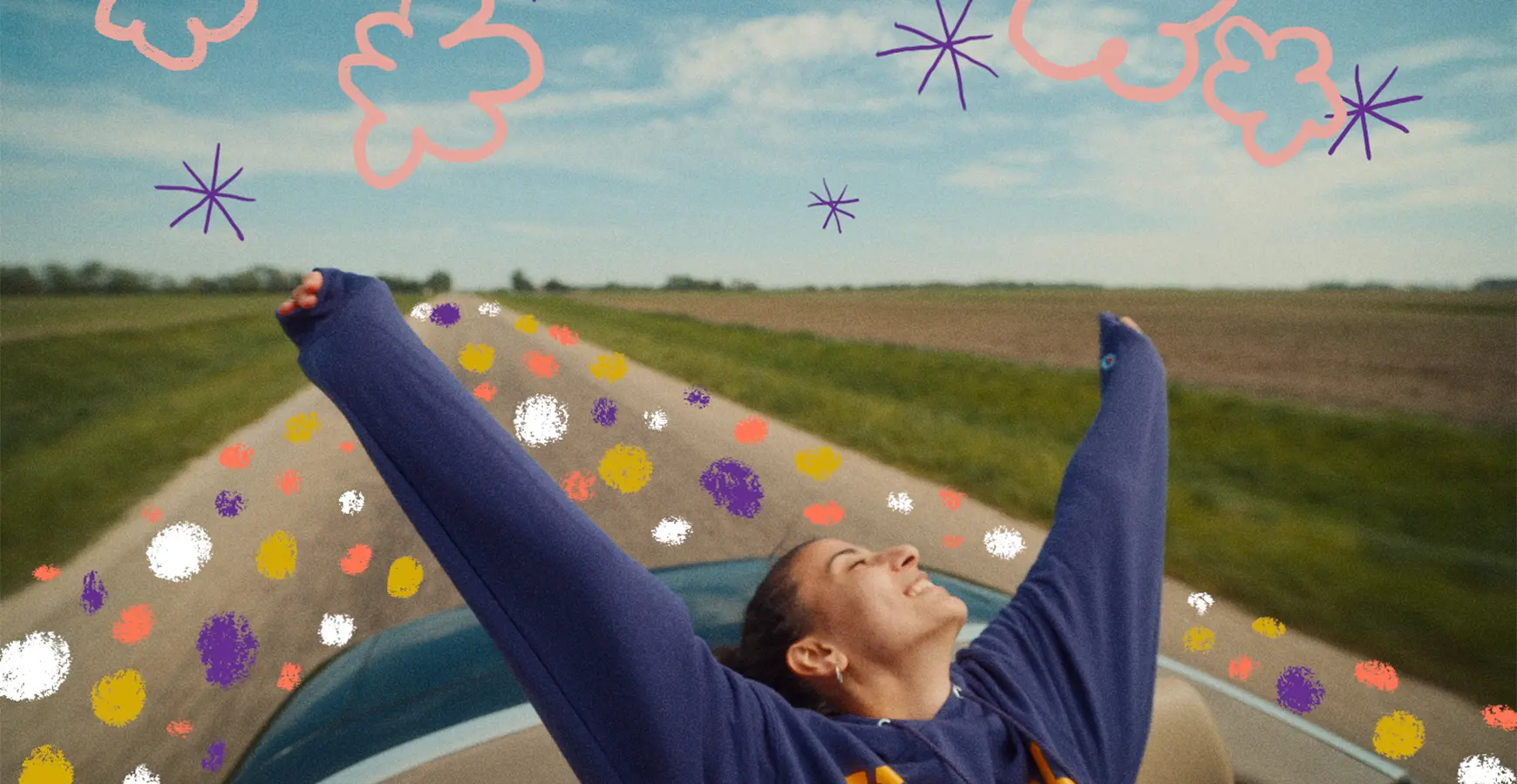 A girl in a convertible outstretches her arms behind her with the road and a prairie behind her.