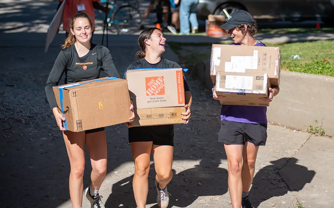 Three KNox students carrying boxes on a campus sidewalk.