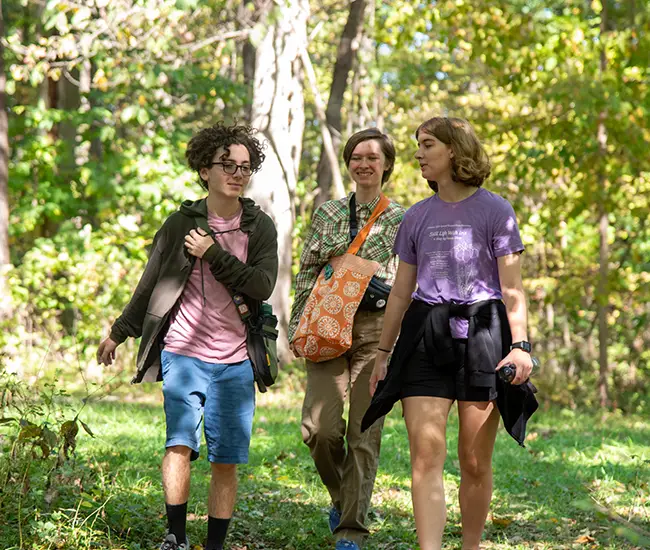 Three students walking outside surrounded by tall green trees and grass.