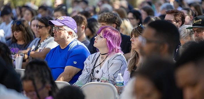 A large group of students sits in an audience outside.