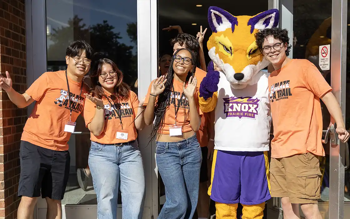 A group of students stand at a door waving at the camera with the mascot Blaze with them.