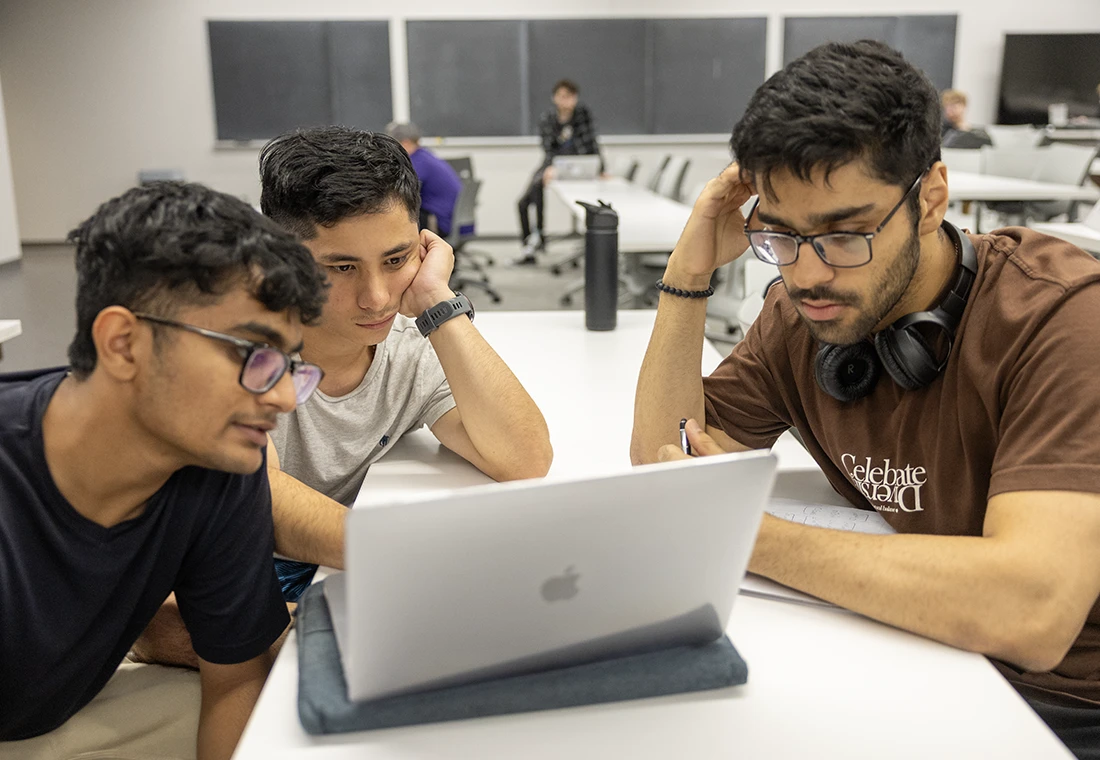 A group of three students sitting at a desk looking at a laptop.
