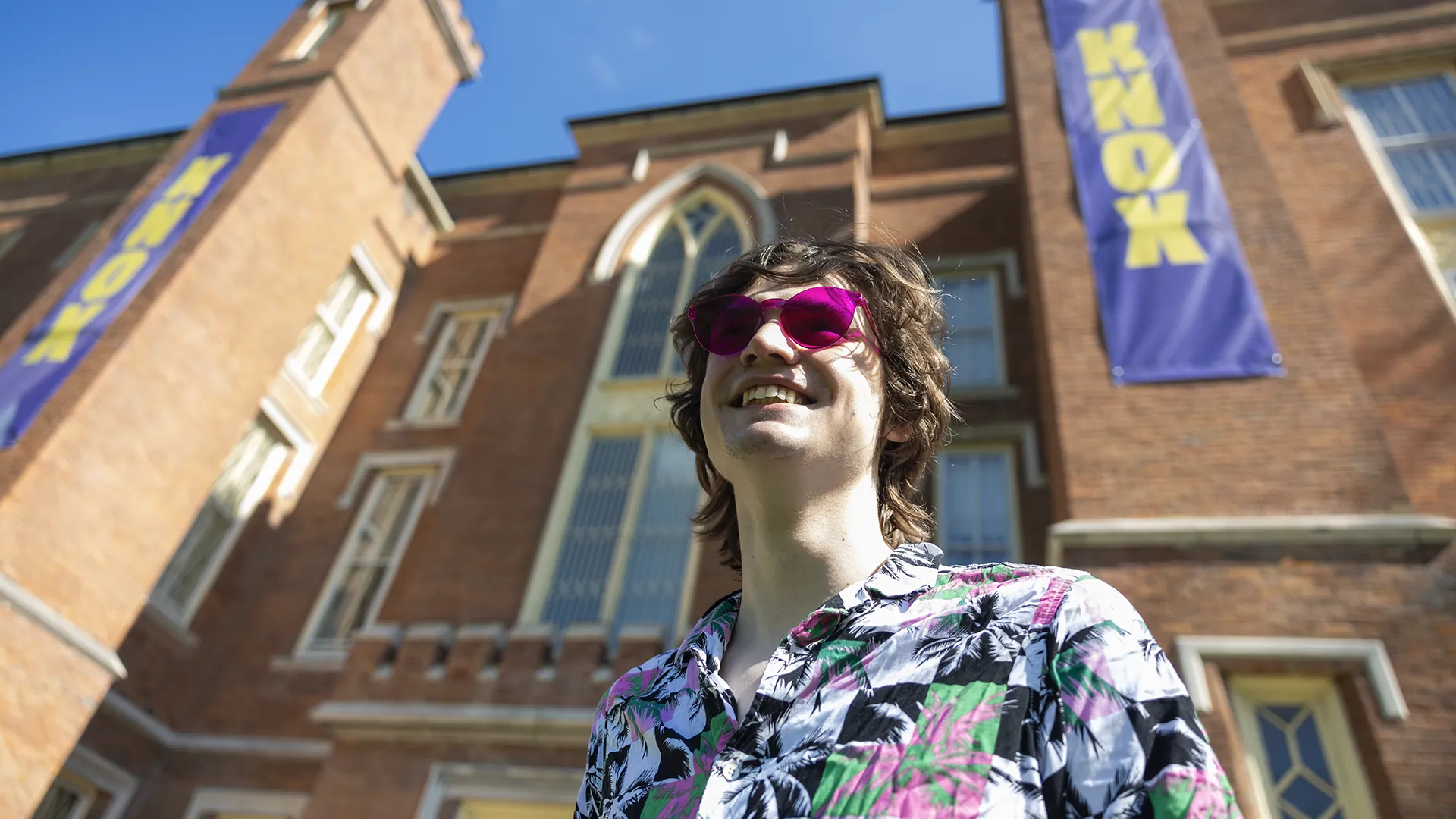 A photo of a smiling student in sunglasses taken from below, with Old Main in the backgroun. A purple banner with yellow letters that spell Knox is hanging on the side of the building.