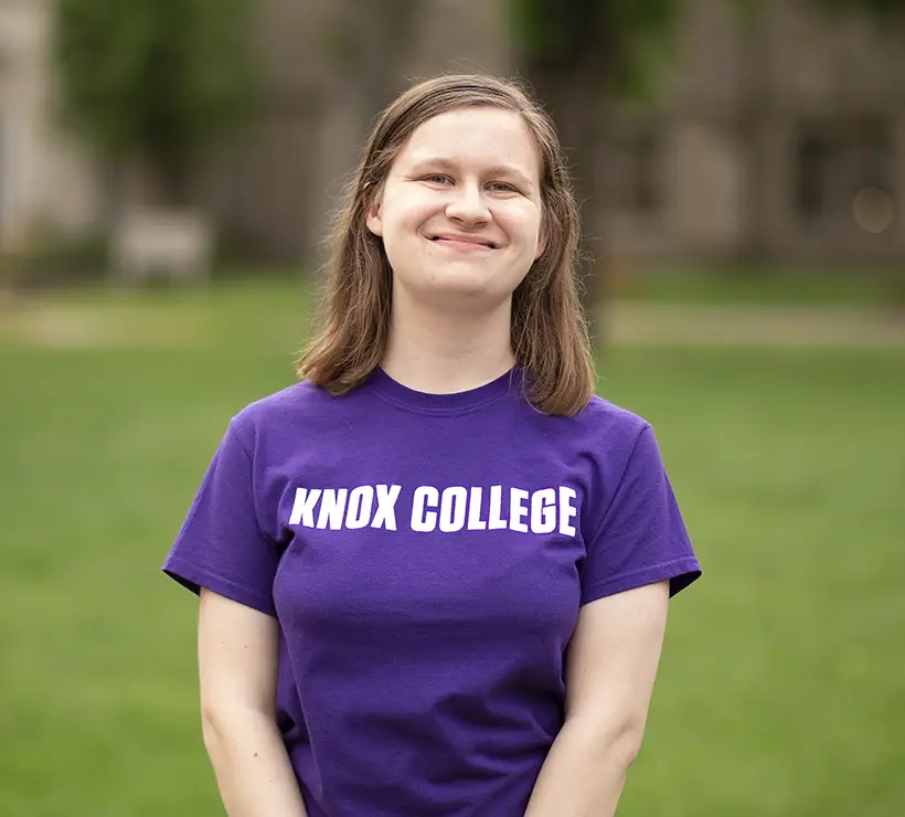 Sarah Wallenfelsz smiling at the camera. She is wearing a purple shirt with white letters that spell Knox College.