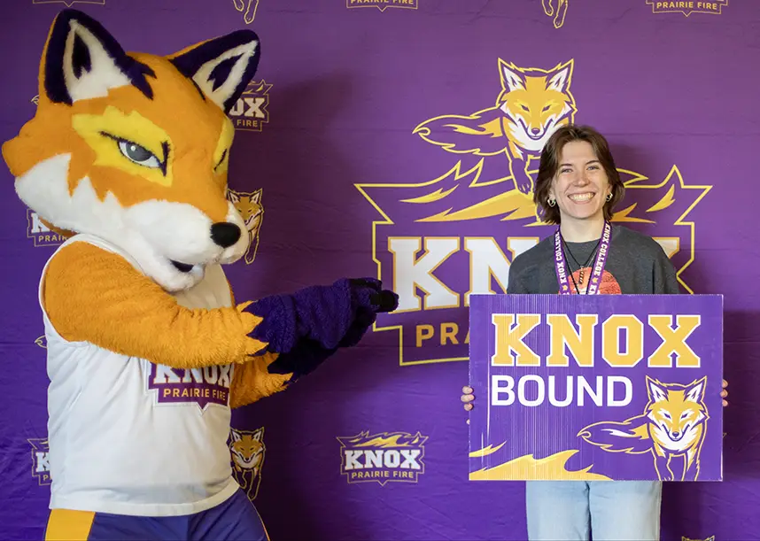 A student stands in front of a Knox step and repeat holding a sign that says "Knox Bound" with Blaze the Knox Fox standing next to them.
