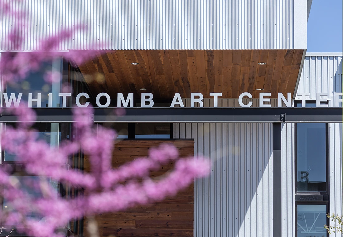 A photo of the Whitcomb Art Center, a large uilding with corregated stell siding and wooden accents, with a pink flowering tree in the forefront.