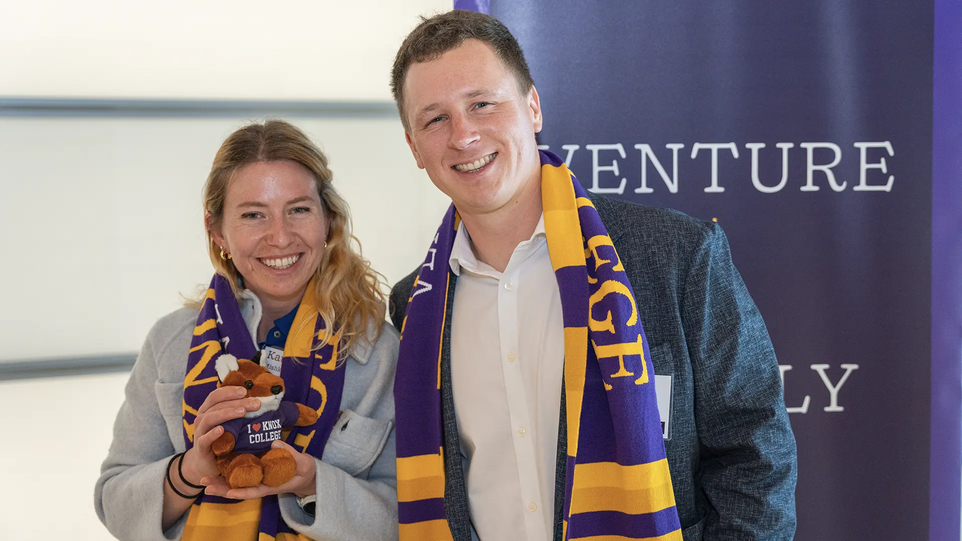 Two alumni smile at the camera in front of a purple banner that says Venture Boldlly in white. They both wear purple and gold scarves, and one hols a stuffed bear with a purple shirt that says, "I heart Knox College."