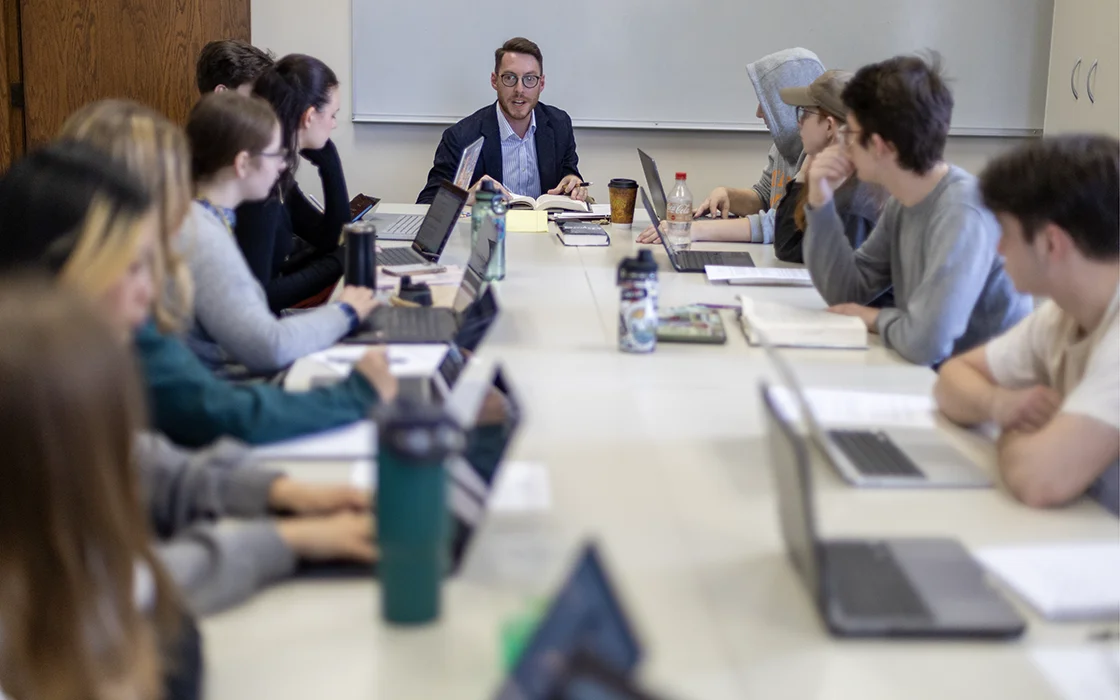 Students with laptops sit around a long table as a Knox professor lectures from the head of the table.