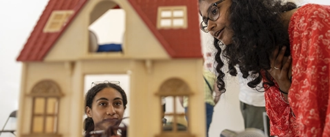 Two Knox students look at a dollhouse, one student is looking from above, while the other is peering in through a window.